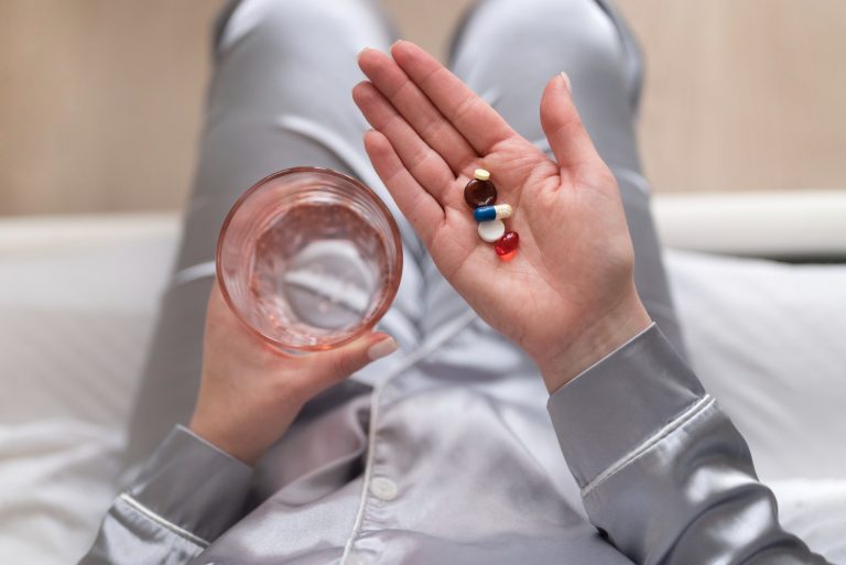 실크 파자마를 입은 여성이 침대 위에 앉아 한 손에는 물이 담긴 유리컵을, 다른 손 손바닥에는 색깔이 다른 여러 알의 영양제 캡슐과 정제를 올려놓고 있는 모습. 갱년기 건강 관리를 위해 복수의 영양제를 함께 복용하는 상황.