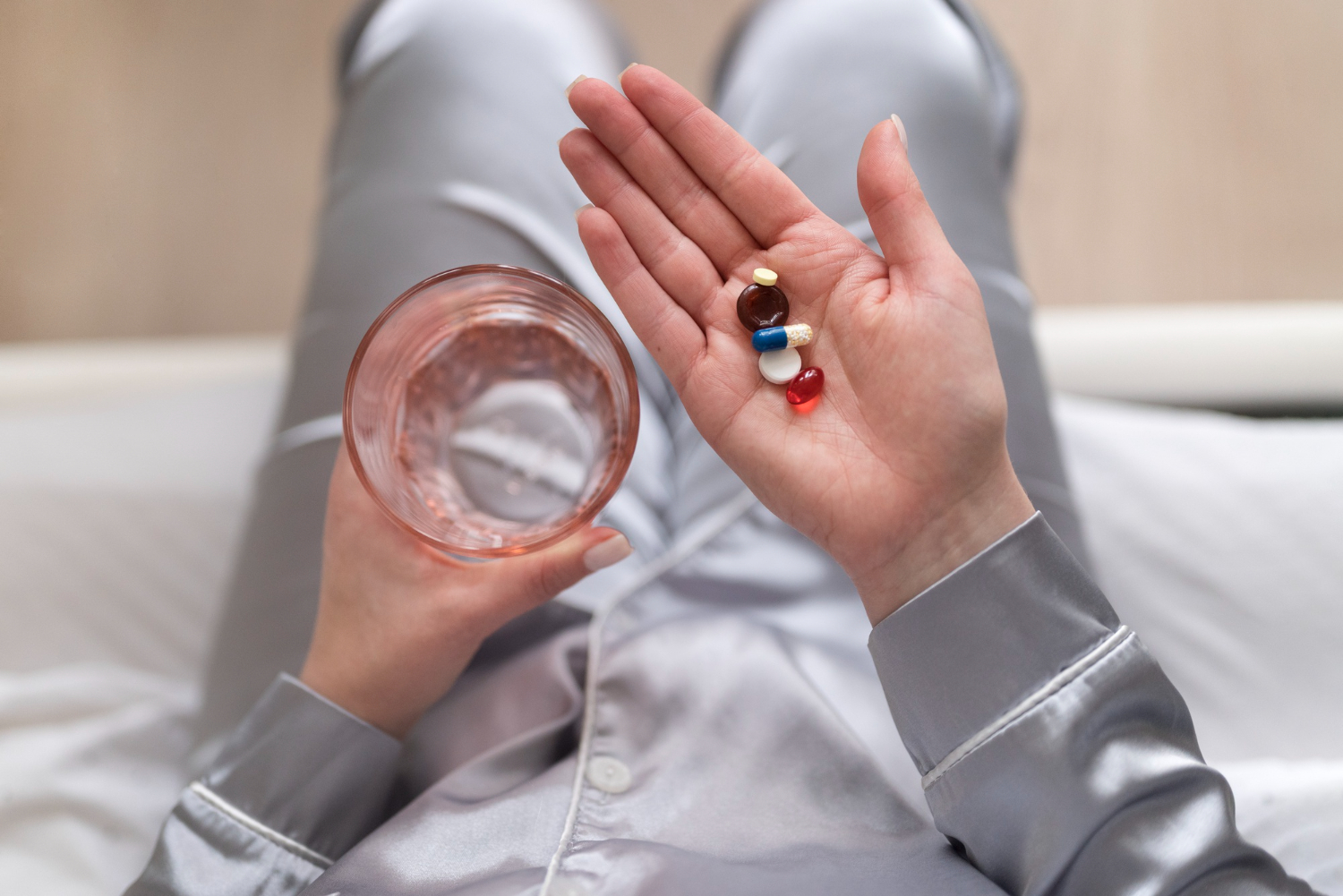 실크 파자마를 입은 여성이 침대 위에 앉아 한 손에는 물이 담긴 유리컵을, 다른 손 손바닥에는 색깔이 다른 여러 알의 영양제 캡슐과 정제를 올려놓고 있는 모습. 갱년기 건강 관리를 위해 복수의 영양제를 함께 복용하는 상황.