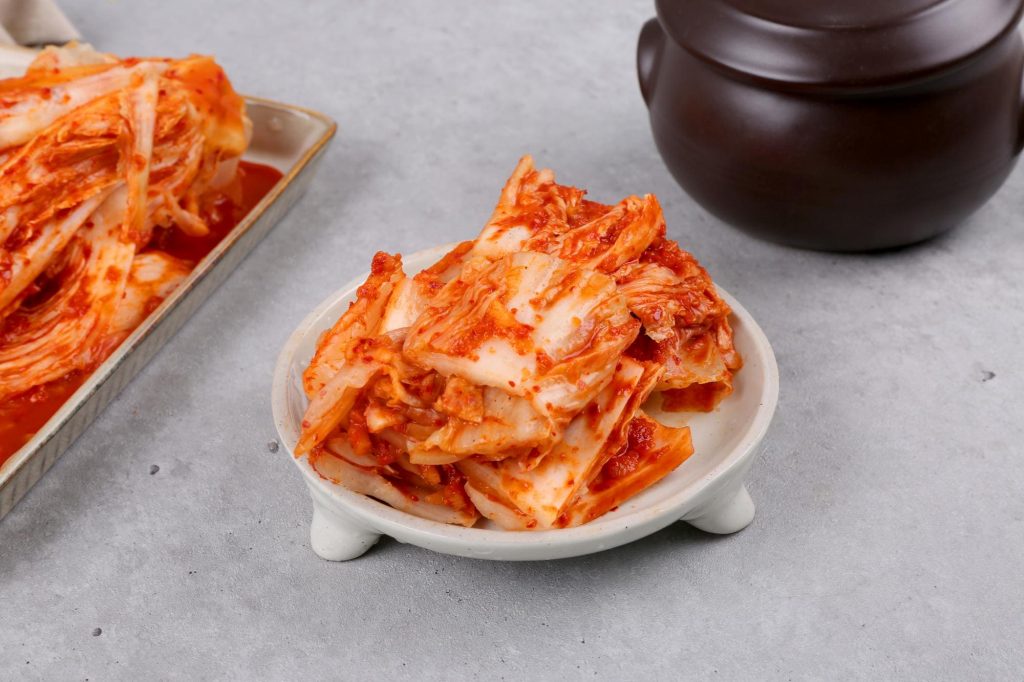 흰 접시에 정갈하게 담긴 빨간 양념의 배추김치와 그 뒤로 보이는 전통 옹기 항아리.