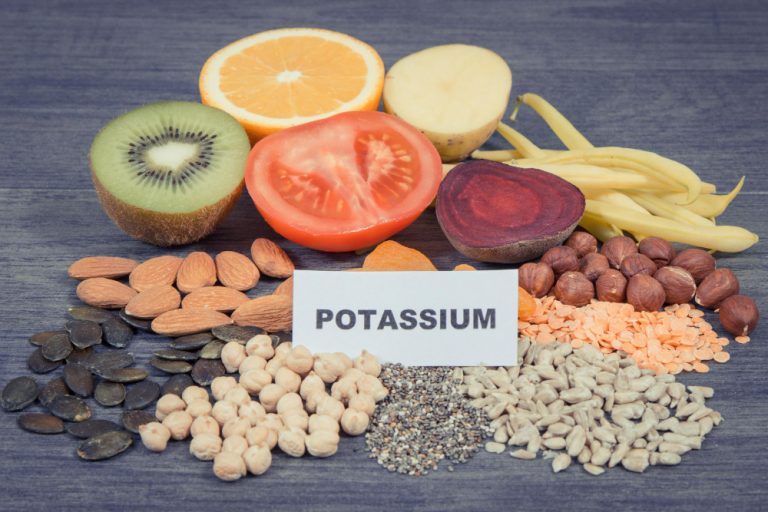 POTASSIUM 표지판과 키위·토마토·콩류·견과류 등 칼륨 풍부 식품 — 채식 식단 고칼륨혈증 원인 식품 종류 Potassium label with kiwi tomato legumes nuts and seeds – high potassium plant foods causing hyperkalemia in vegan vegetarian diet