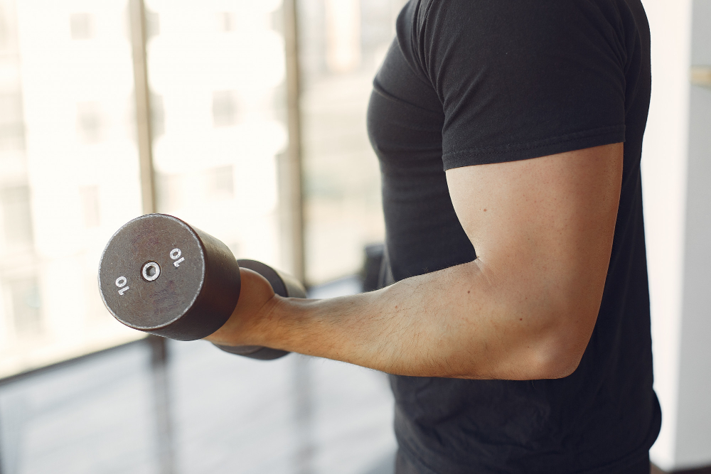 검은 티셔츠를 입은 남성이 창가에서 덤벨 컬 운동을 하는 모습. 규칙적인 저항성 운동은 근육 단백질 합성을 촉진하고 근감소 진행을 늦추는 데 기여한다.